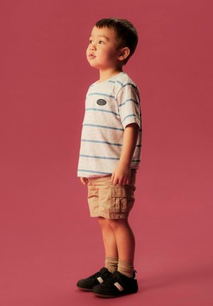 Junger Junge steht mit dem Gesicht nach links, trägt ein gestreiftes T-Shirt, beige Cargoshorts, beige Socken und schwarze Sneakers vor einem einfachen rötlichen Hintergrund.