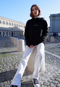 Felpa nera con testo bianco, jeans bianchi larghi e sneaker nere, seduto su un blocco di pietra in un'area lastricata di ciottoli, la luce del sole illumina la scena.