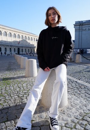 Felpa nera con testo bianco, jeans bianchi larghi e sneaker nere, seduto su un blocco di pietra in un'area lastricata di ciottoli, la luce del sole illumina la scena.