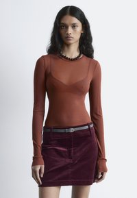 Langärmliges, durchsichtiges rotes Oberteil kombiniert mit einem tief burgunderfarbenen Cordrock. Verfügt über einen grauen Gürtel und eine runde, dunkle Perlenkette. Insgesamt glatte Textur.