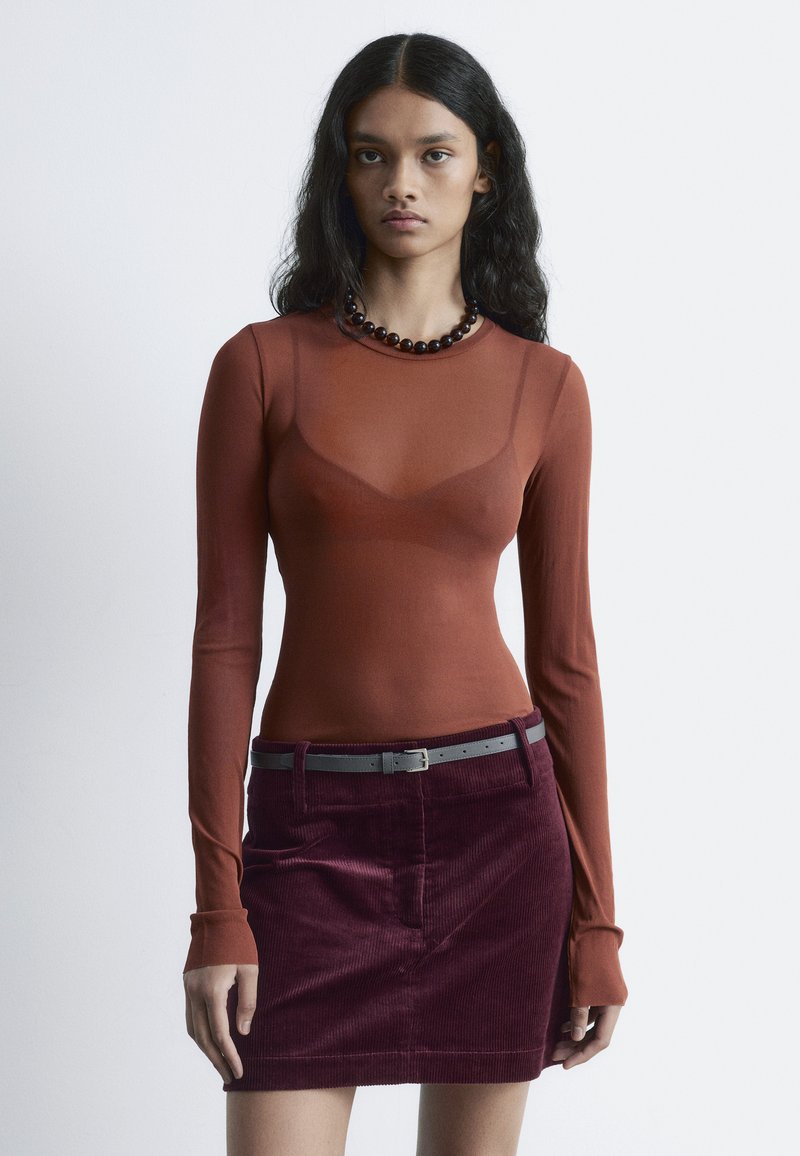 Langärmliges, durchsichtiges rotes Oberteil kombiniert mit einem tief burgunderfarbenen Cordrock. Verfügt über einen grauen Gürtel und eine runde, dunkle Perlenkette. Insgesamt glatte Textur.