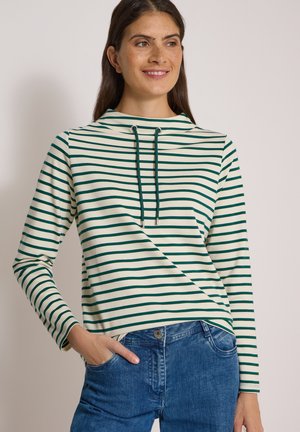 Femme portant un haut à manches longues rayé blanc et vert avec des cordons, souriante, et un jean en denim bleu, main dans la poche, sur un fond uni.