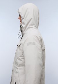 Parka gris claro con capucha, que cuenta con cordones ajustables, cierre frontal con cremallera, grandes bolsillos y un parche con el logo en el hombro.