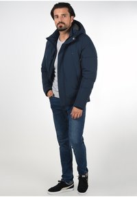 Marineblå isoleret jakke med hætte, lynlås foran og to sidelommer. Bæres over en grå skjorte og parret med blå jeans og sorte sneakers.