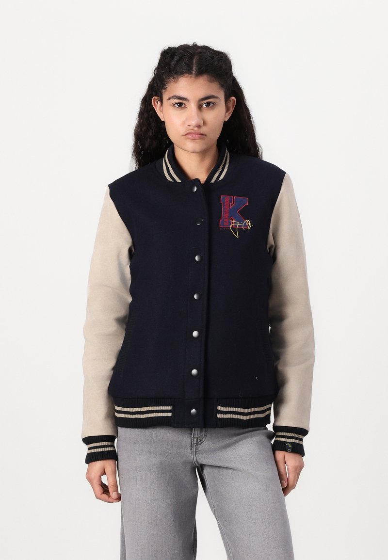 Marineblaue Varsity-Jacke mit beigen Ärmeln, gestreiftem, geripptem Kragen und Bündchen, vorderen Druckknöpfen und gesticktem Logo auf der Brust.