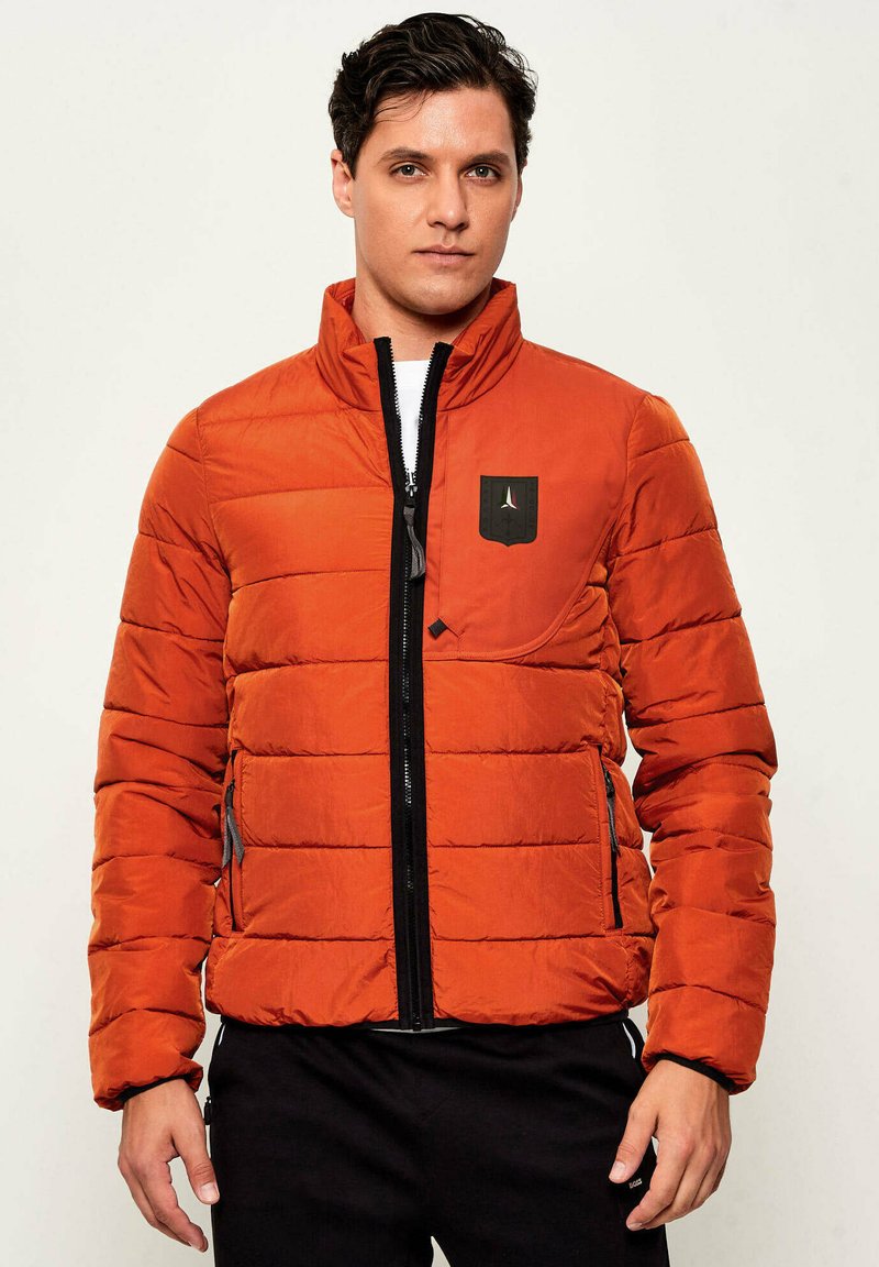 Giacca imbottita arancione con colletto rialzato, zip frontale e patch con logo nero. Presenta due tasche laterali e una trama trapuntata.