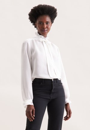 Weißes Langarmhemd mit hohem Rüschensaum, vertikalen Streifen und enganliegenden Manschetten, kombiniert mit schwarzen High-Waist-Jeans.