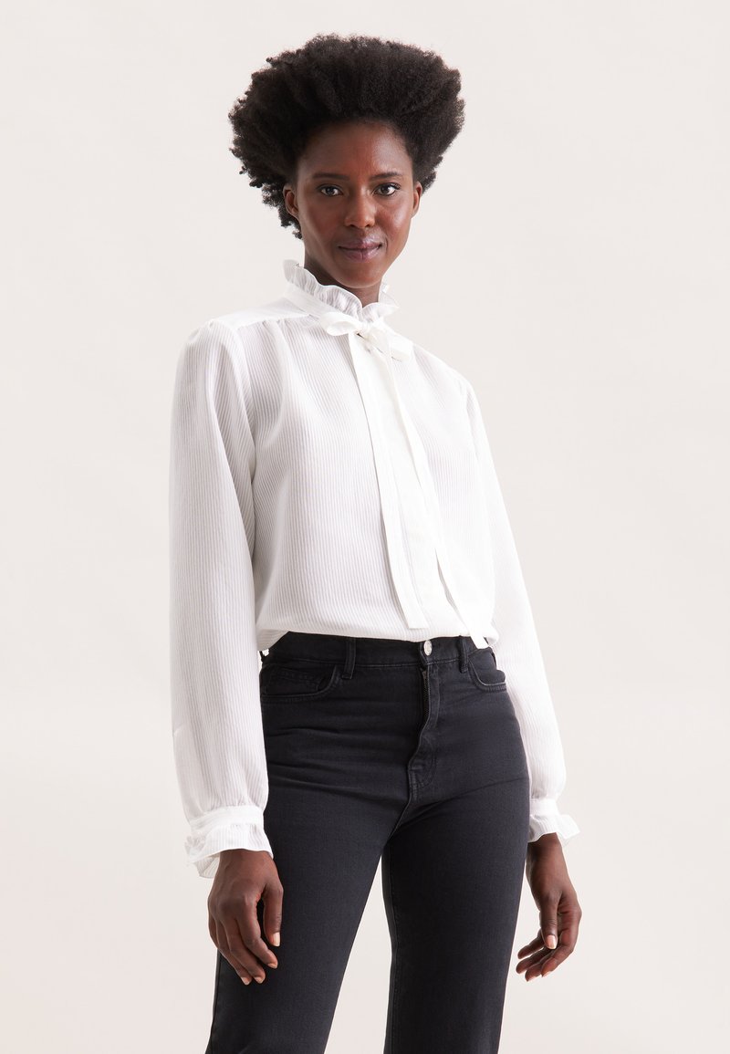 Weißes Langarmhemd mit hohem Rüschensaum, vertikalen Streifen und enganliegenden Manschetten, kombiniert mit schwarzen High-Waist-Jeans.