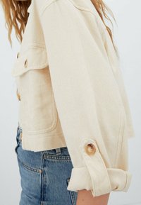 Jasno beżowa, skrócona kurtka dżinsowa z dwoma kieszeniami na piersi, rękawami z możliwością podwinięcia oraz jasnobrązowymi guzikami, prezentująca teksturowany materiał i swobodny fason.