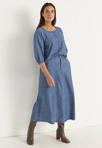 Jeansklänning med trekvartslång ärm, rund halsringning och midja med bälte. Har en utsvängd kjol och subtila sömnadsdetaljer. Bruna klackstövlar.