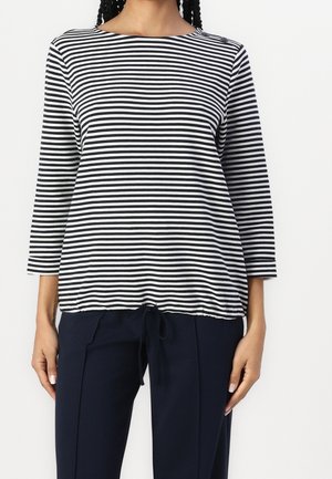 Femme portant un t-shirt à manches longues à rayures horizontales noir et blanc avec un pantalon bleu marine, debout sur un fond blanc.