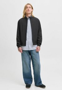 Zwarte bomberjack met rits, ribgebreide manchetten, gedragen over een blauw gestreept overhemd met knopen, in combinatie met wijde denim jeans.