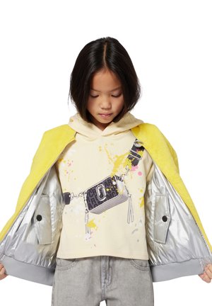 Kind met schouderlang donker haar draagt een gele jas die open is, en laat een crèmekleurige hoodie zien met een crossbody tas en een verfspatontwerp.