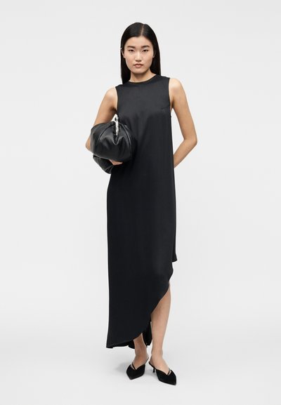 Vestido preto sem mangas com bainha assimétrica, tecido suave e design minimalista. A modelo segura uma mala redonda preta e usa sapatilhas pontiagudas.