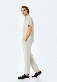 Camicia corta a maniche corte beige a coste, pantaloni beige chiaro e sneaker nere con lacci bianchi. Il modello è in posa di profilo con le mani in tasca.