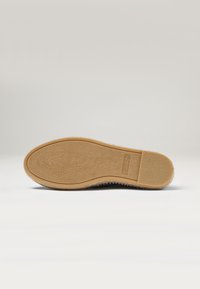 Suela de un zapato con una base de goma marrón texturizada y un contorno liso; incluye una etiqueta de marca cerca del talón.