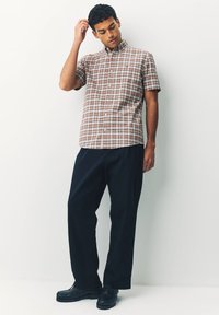 Chemise à manches courtes, à carreaux marron, avec col boutonné, associée à un pantalon ample foncé et des chaussures noires.