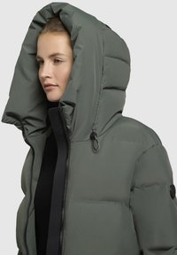 Donkergroene pufferjack met een grote capuchon, ritssluitingen en zwarte accenten. Het heeft een gequiltde textuur en een verstelbaar koord in de capuchon.