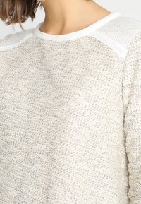 Světle béžový pletený svetr s texturovaným vzorem. Obsahuje bílé akcenty na ramenou a kulatý výstřih. Měkký materiál, volný střih.