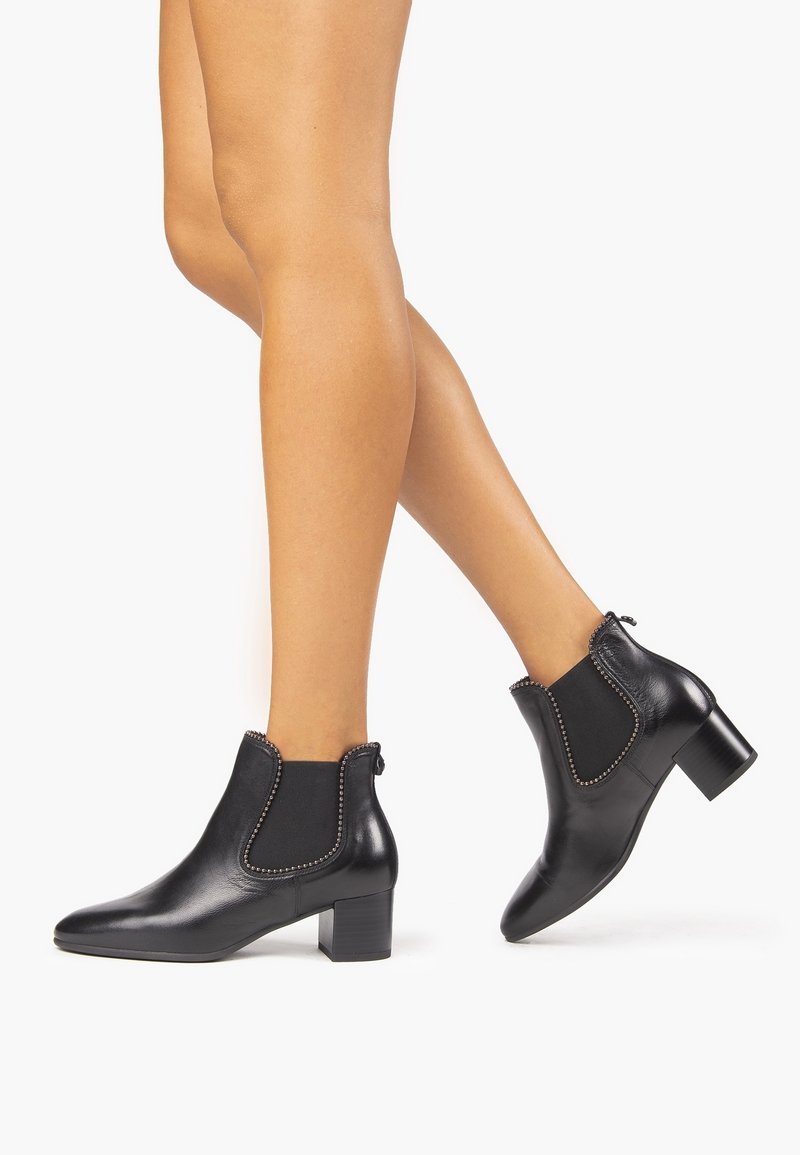 Jambes portant des bottines en cuir noir avec talons carrés et bordure cloutée, debout sur un fond blanc.