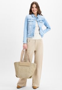 Beige stoffen tote bag met een gestructureerde weving, een effen tan accent en handvatten van gemiddelde lengte. Model draagt een lichtblauwe denimjas en beige broek.