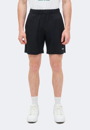 Muž oblečený v čiernych športových šortkách, bielych ponožkách a bielych teniskách stojaci pred jednoduchým svetlým pozadím.