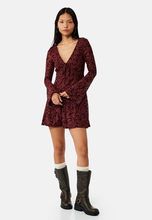 Kvinde iført en burgunderfarvet blomsterprintet langærmet romper med klokkeærmer og sorte midt-lægsstøvler med hvide sokker mod en ensfarvet baggrund.