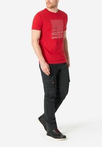 Rotes Baumwoll-T-Shirt mit weißem grafischem Textdesign, kombiniert mit schwarzen Cargo-Hosen und schwarzen Schuhen mit roten Akzenten und weißen Sohlen.