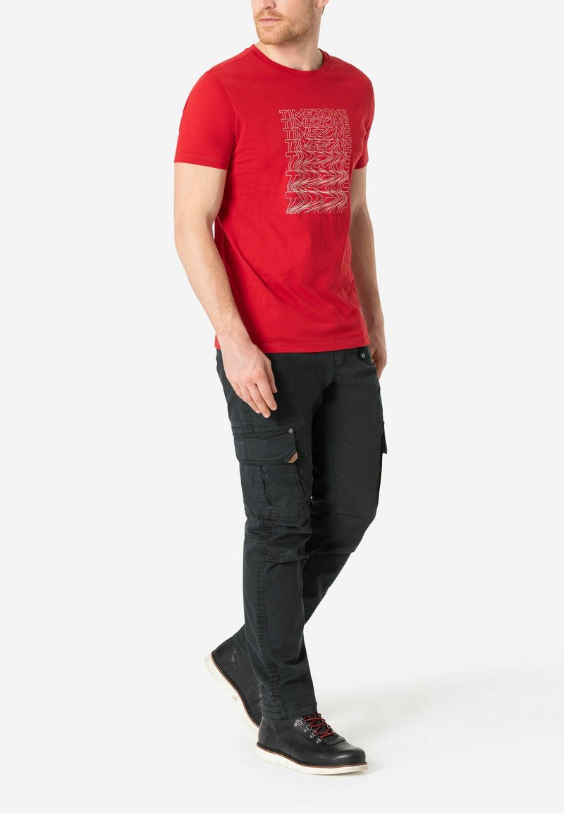 Rotes Baumwoll-T-Shirt mit weißem grafischem Textdesign, kombiniert mit schwarzen Cargo-Hosen und schwarzen Schuhen mit roten Akzenten und weißen Sohlen.