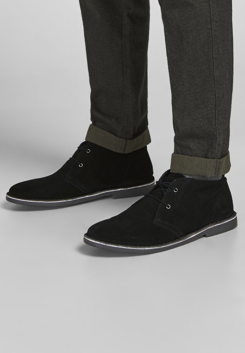 Personne portant des bottines noires en daim à lacets et un pantalon foncé retroussé sur un sol gris clair.