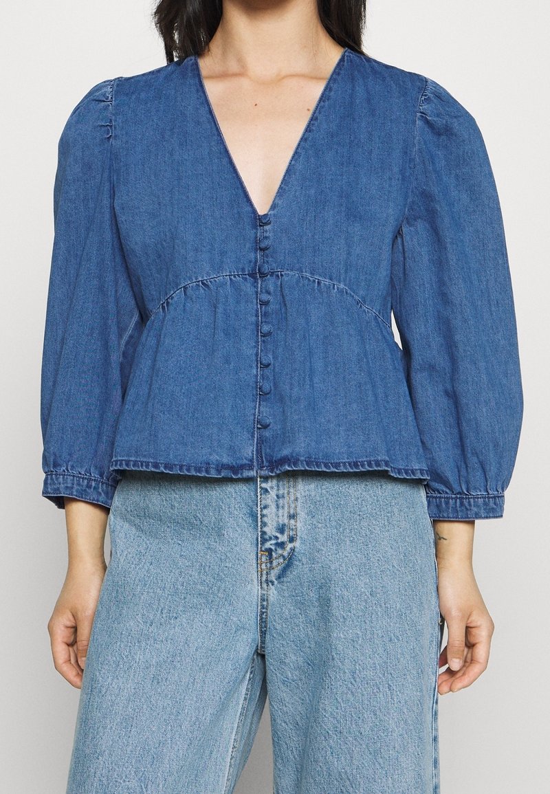 Denim peplum top s hlubokým V-výstřihem, dlouhými puff rukávy a centrálním zapínáním na knoflíky. Světle modrá, s nařaseným pasem a volným střihem.