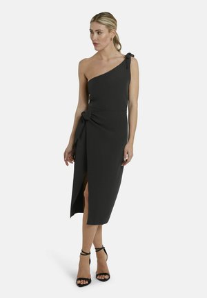 Vrouw die een zwarte, asymmetrische jurk tot op de knie draagt met een zijsplitsing en strikdetails, gecombineerd met zwarte hoge hakken met enkelbandje.