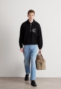 Mustaga tõmbatav must kott, millel on logo, helesinised teksad ja mustad jalanõud, kandes heledate aktsentidega pruuni kotti. Sile kangas ja mugav lõige.