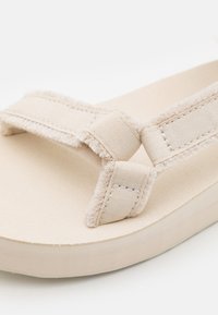 Beige sandal with soft fabric straps, textured finish, and a cushioned sole. Features wide straps and a flat, minimalistic design.