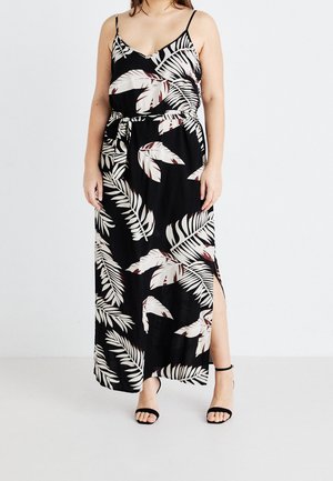Frau trägt ein schwarzes, ärmelloses Maxikleid mit weißem und braunem tropischem Blattmuster und seitlichem Schlitz, kombiniert mit schwarzen Absatzschuhen mit Knöchelriemen.