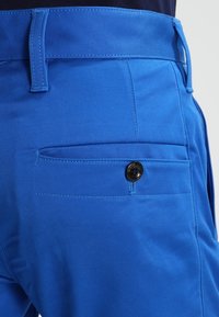 Modré kalhoty s hladkou strukturou, s kapsou na zadní straně zabezpečenou černým knoflíkem. Švy podél pasu.