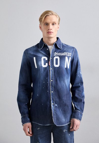 Chemise en denim bleu foncé avec du texte blanc "ICON". Elle se ferme par des boutons sur le devant, possède un col pointu et des accents usés avec des éclaboussures de peinture.