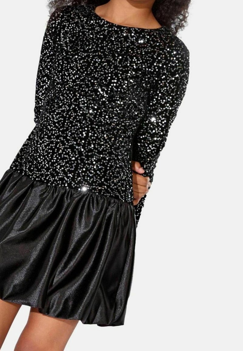 Enfant portant une robe noire à manches longues avec des sequins scintillants sur le haut et une jupe brillante froncée, debout devant un fond uni.