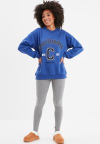 Sudadera azul con el texto "California", leggings grises y zapatillas de casa de color marrón con acentos de piel blanca. La modelo posa con las manos en las caderas.