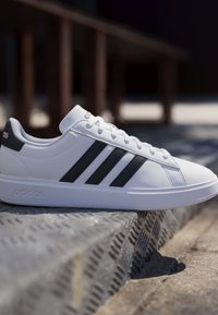 Weißer Adidas-Sneaker mit schwarzen Streifen, der auf einer strukturierten Metallstufe im Freien bei Tageslicht ruht.