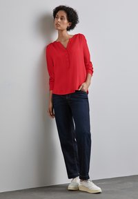 Röd långärmad blus med V-ringning och knappslå, kombinerad med mörkblå jeans och vita sneakers. Slät textur, avslappnad passform.