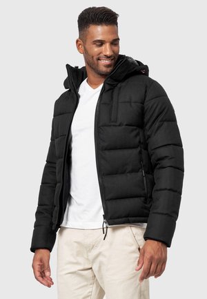 Chaqueta de plumas negra con capucha, con cierre de cremallera, puños elásticos y un acabado texturizado. Llevada sobre una camisa blanca y pantalones claros.