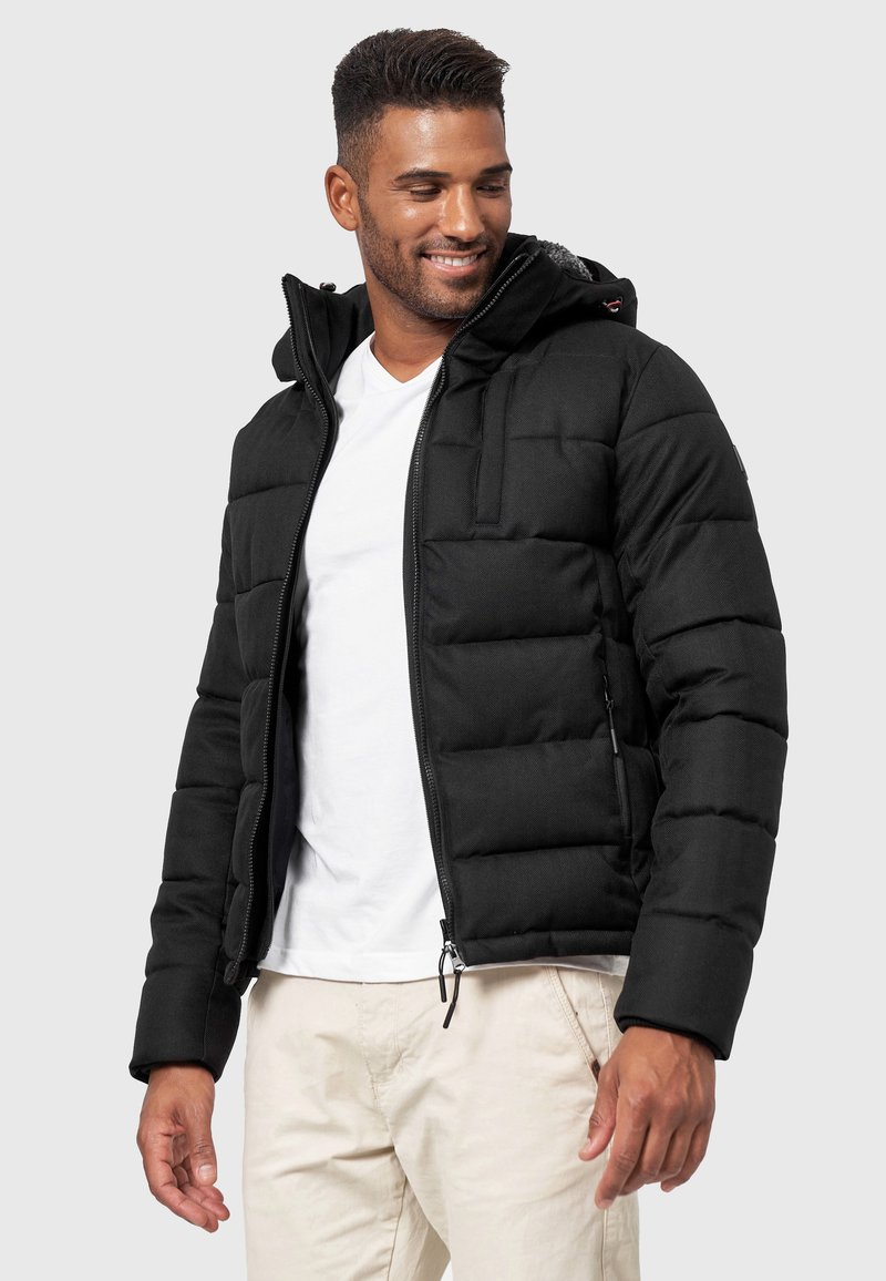 Schwarze Puffjacke mit Kapuze, ausgestattet mit einem Reißverschluss, elastischen Bündchen und einer strukturierten Oberfläche. Getragen über einem weißen Hemd mit hellen Hosen.