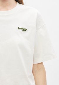 Camiseta blanca de algodón con mangas cortas que presenta un logo bordado verde de Lacoste en el lado izquierdo del pecho. Diseño liso, ajuste estándar.