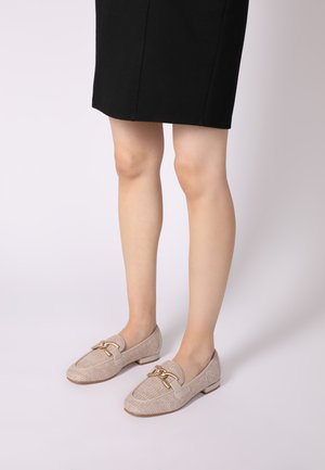 Beige stof loafers met een textuurafwerking, platte hak en een gouden kettingaccent aan de voorkant, gecombineerd met een zwarte rok.