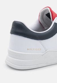 Sneaker bianco con tacco in pelle blu navy, marchio dorato "HILFIGER", suola bianca testurizzata e imbottitura interna rossa. Design liscio e strutturato.