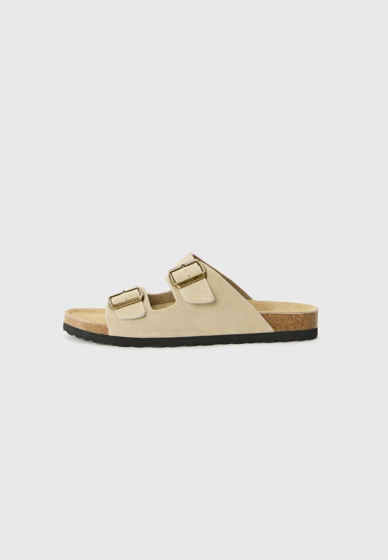 Beige Wildleder-Open-Toe-Slide-Sandale mit zwei verstellbaren goldfarbenen Schnallenriemen und Korkfußbett auf schwarzer Gummisohle.