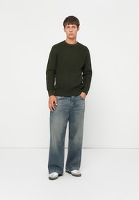 Maglione a maglia verde scuro con un motivo texturizzato, abbinato a jeans azzurri larghi. Il modello è in piedi contro uno sfondo bianco uniforme.