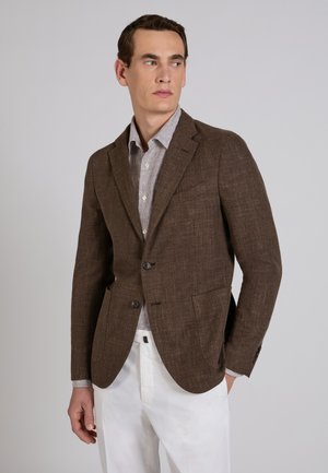 Mężczyzna w brązowej marynarce na paskowanej koszuli i białych spodniach, stojący z jedną ręką w kieszeni, patrzący na bok.