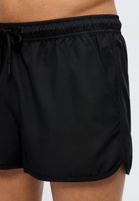 Κοντινή λήψη μαύρου σορτς με ελαστική μέση, πλαϊνή τσέπη και κορδόνι, φορεμένο από άτομο με ορατό δέρμα στην περιοχή της μέσης και του μηρού.